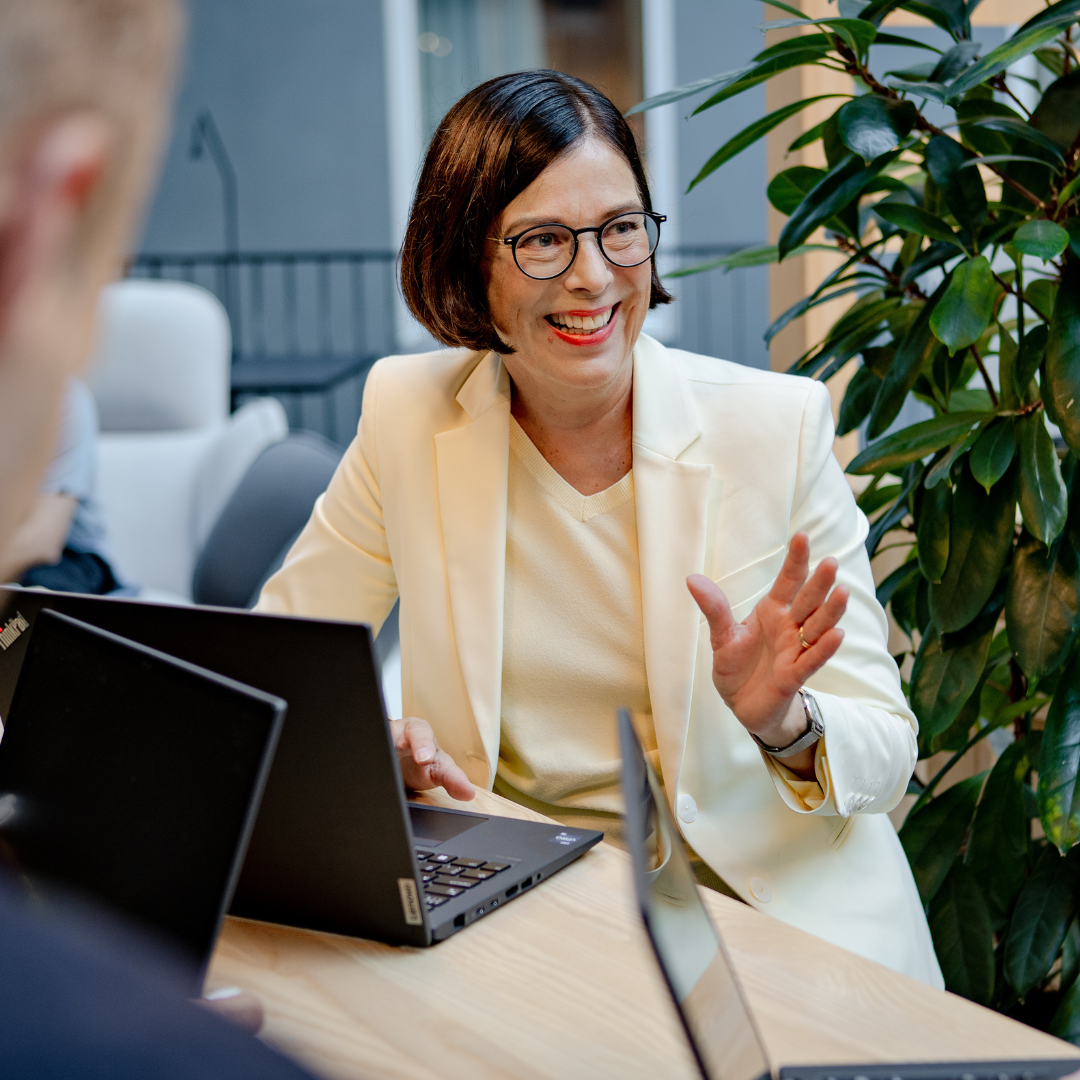 Dame i blaser som smiler ved et bord og gestikulerer mens hun prater