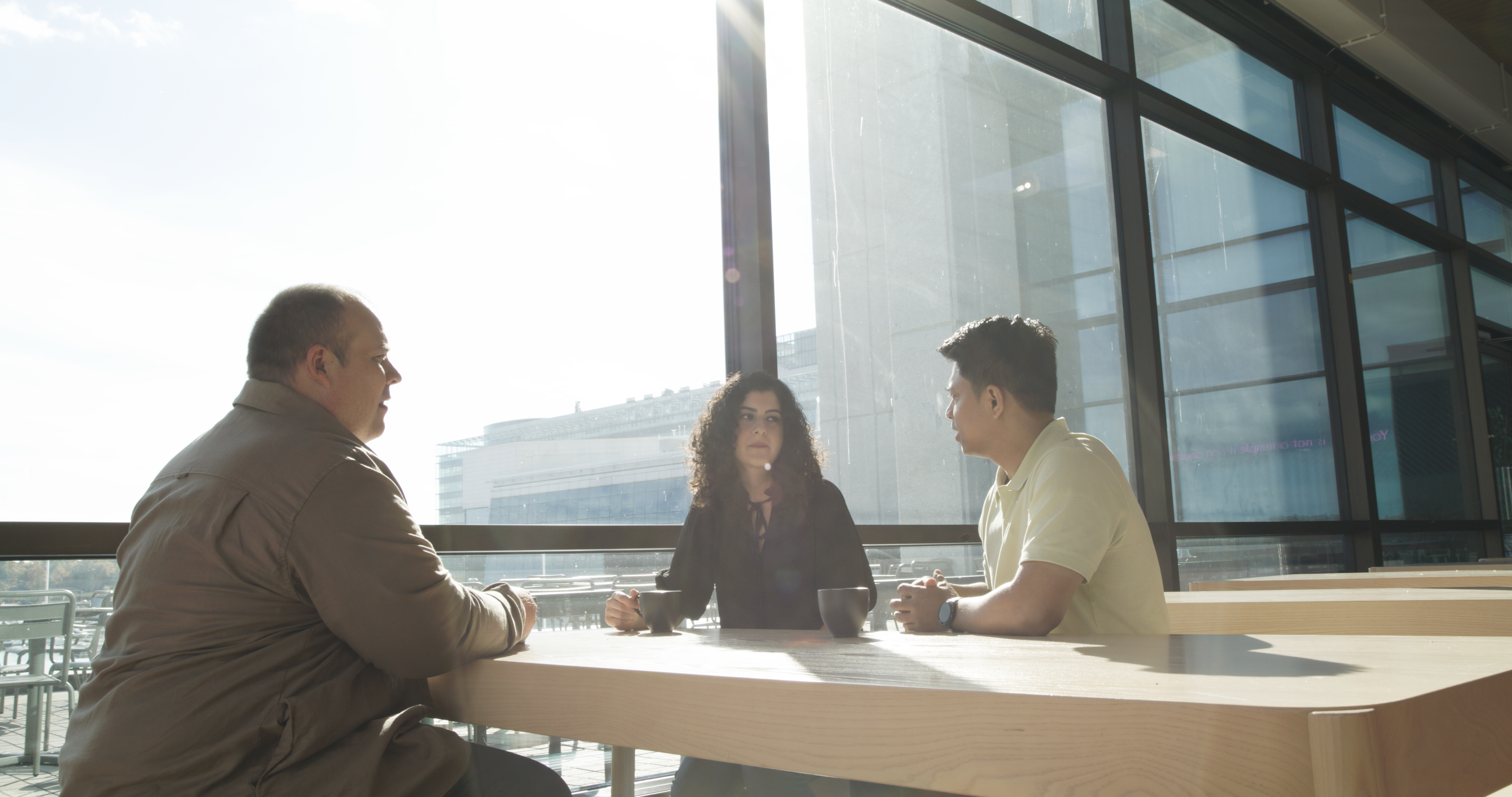 Kollegaer som prater over et bord innendørs i solskinn. Store glassvinduer og et tykt trebord
