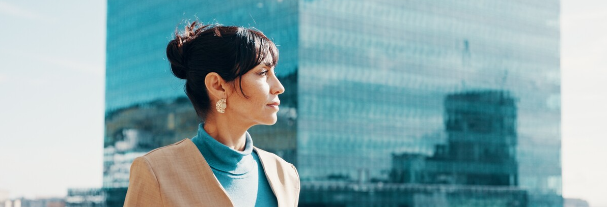 Kvinne som står ute med en skyskraper i bakgrunnen. Hun stirrer utover landskapet. Det er fint vær, muligens litt bris. Hun tenker over neste steg.