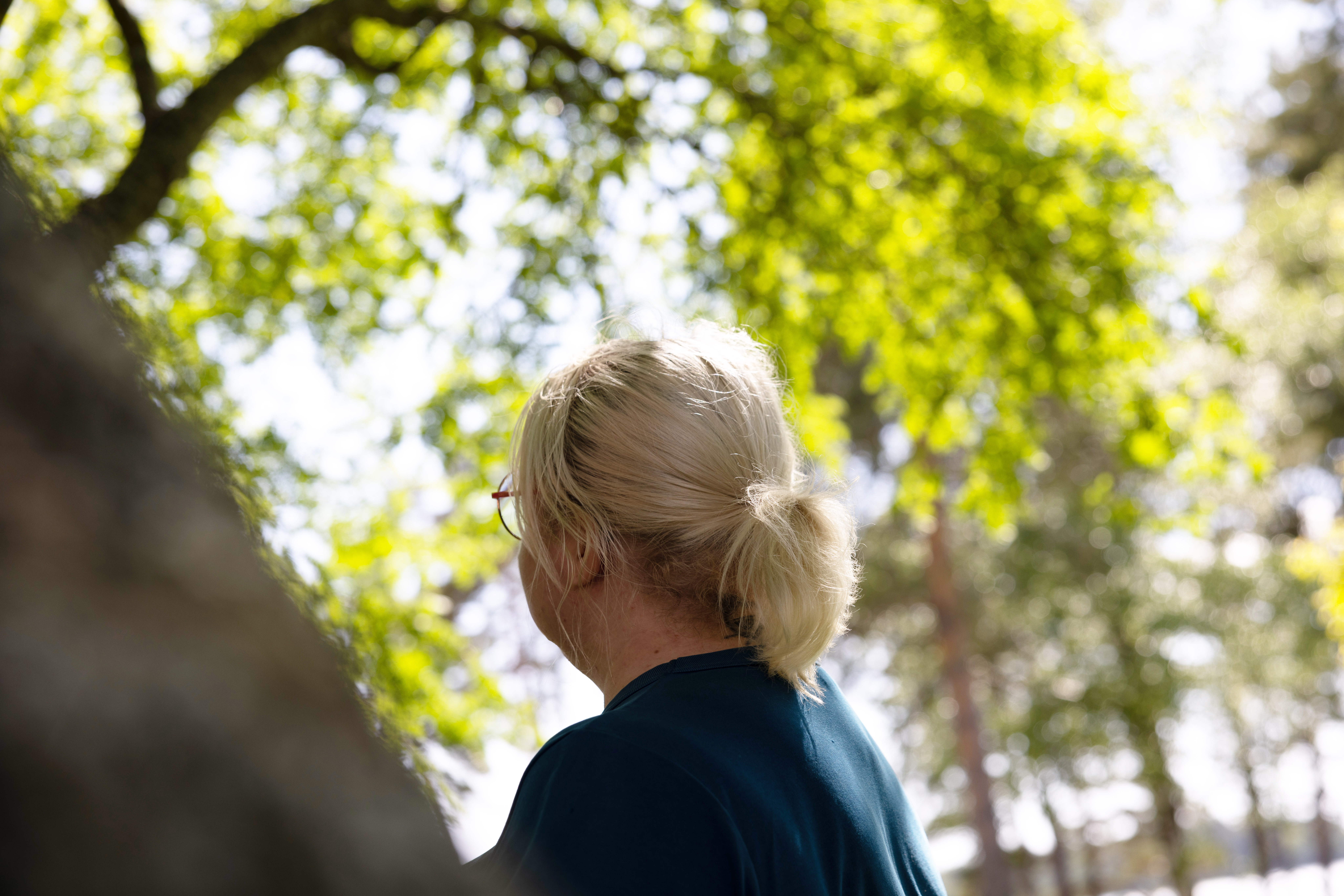 Dame som står ute i solen med et stort grønt tre foran seg som gir litt skygge