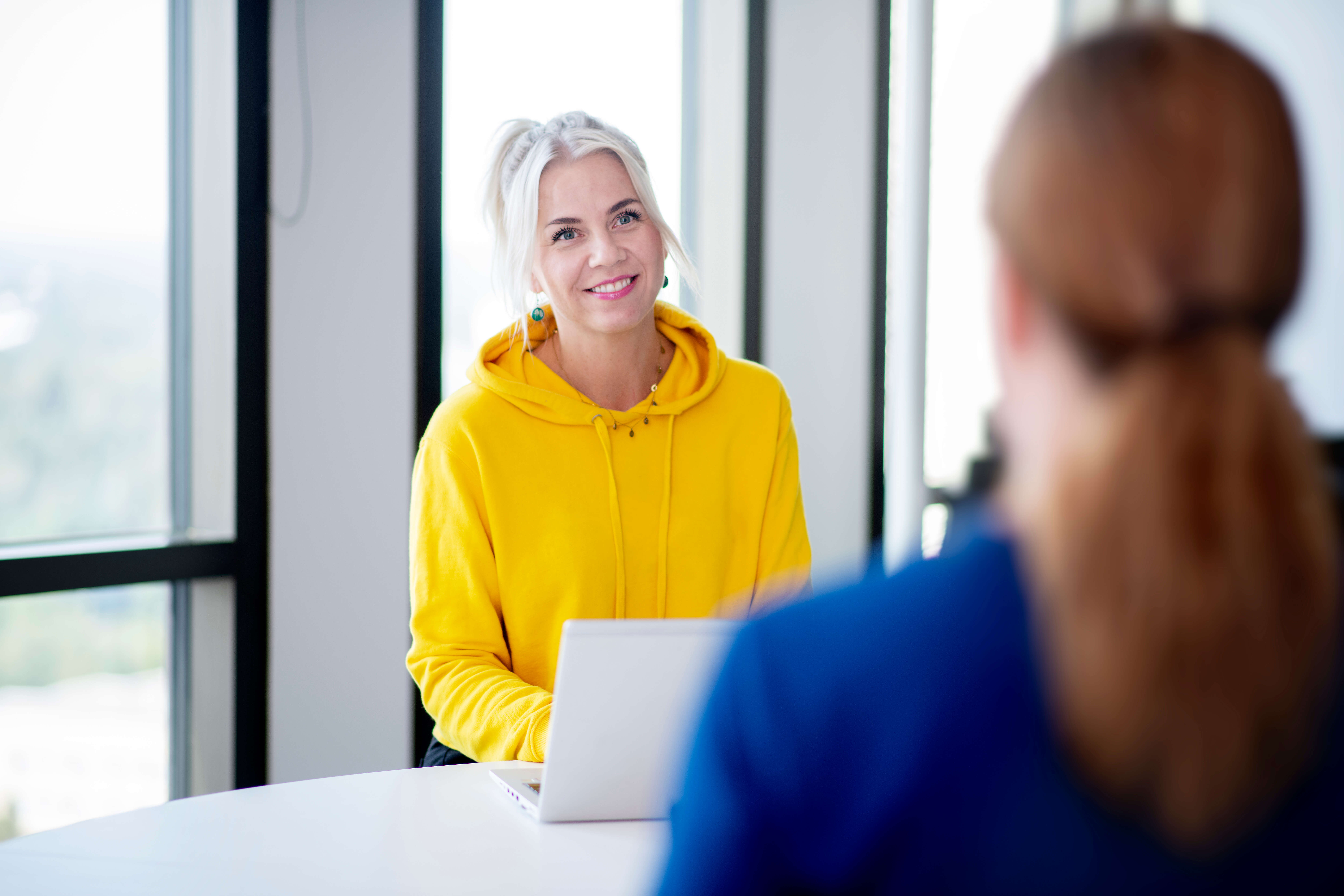 Kontor møte med 2 kvinner som står på hver sin side av et bord. En i gul genser og en i blå genser.