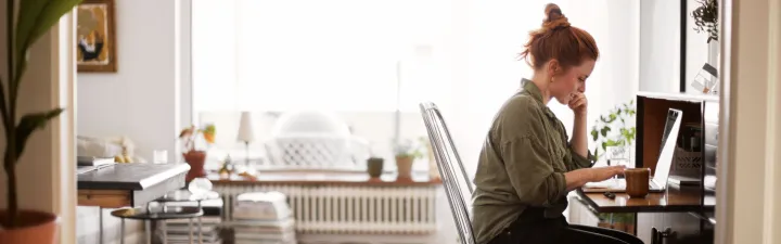 Dame som sitter med pcen sin ved et skrivebord og tenker 