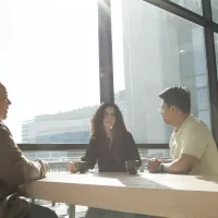 Kollegaer som prater over et bord innendørs i solskinn. Store glassvinduer og et tykt trebord