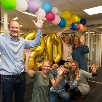 Paul Arne og hans ansatte foran 20 års banneren med fargerike ballonger. Alle har hendene i været og smiler bredt.