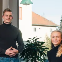 Ung mann og kvinne som står og smiler mot kamera i et kontorlandskap. hun sitter til høyre for han i en stol, mens hans står til venstre. Begge har på seg en svart turtleneck og blå jeans.