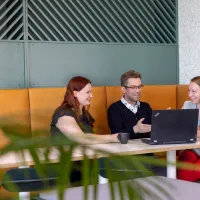 Tre kolleger kolleger i en oransje sofa med grønn vegg og et par kaffekopper