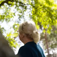 Dame som står ute i solen med et stort grønt tre foran seg som gir litt skygge