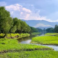 Et fint grønt og frodig område med utsikt til fjellet, en liten bekk som renner igjennom og vakre trær. Det er nesten skyfritt med kun et par små skyer i horisonten