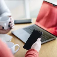To kvinner som sitter sammen over en laptop. den ene holder i en telefon. Man kan kun se armene deres ingen ansikter.